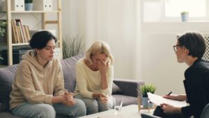 Therapy session in a living room: two clients sit on a sofa, one distressed, as a clinician takes notes nearby.
