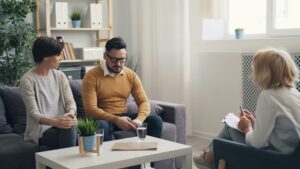 Two people sit on a gray sofa and talk with a clinician taking notes in a bright, cozy office setting.