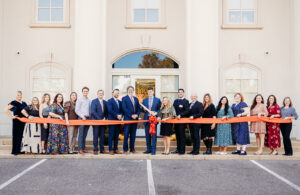 Group of adults standing in a line outdoors for a ribbon-cutting ceremony in front of a beige building with arched windows, holding an orange ribbon.