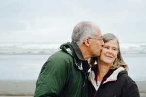 Older couple on a windy beach sharing a kiss on the temple while embracing, with the ocean in the background
