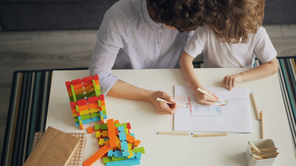 Two children draw on a white table with colorful wooden blocks scattered nearby.