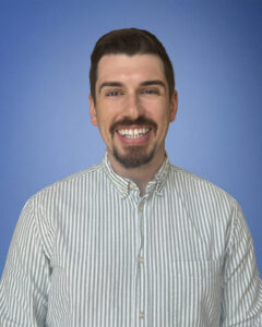 Smiling man with dark hair and a goatee wearing a striped button-up shirt against a blue background.