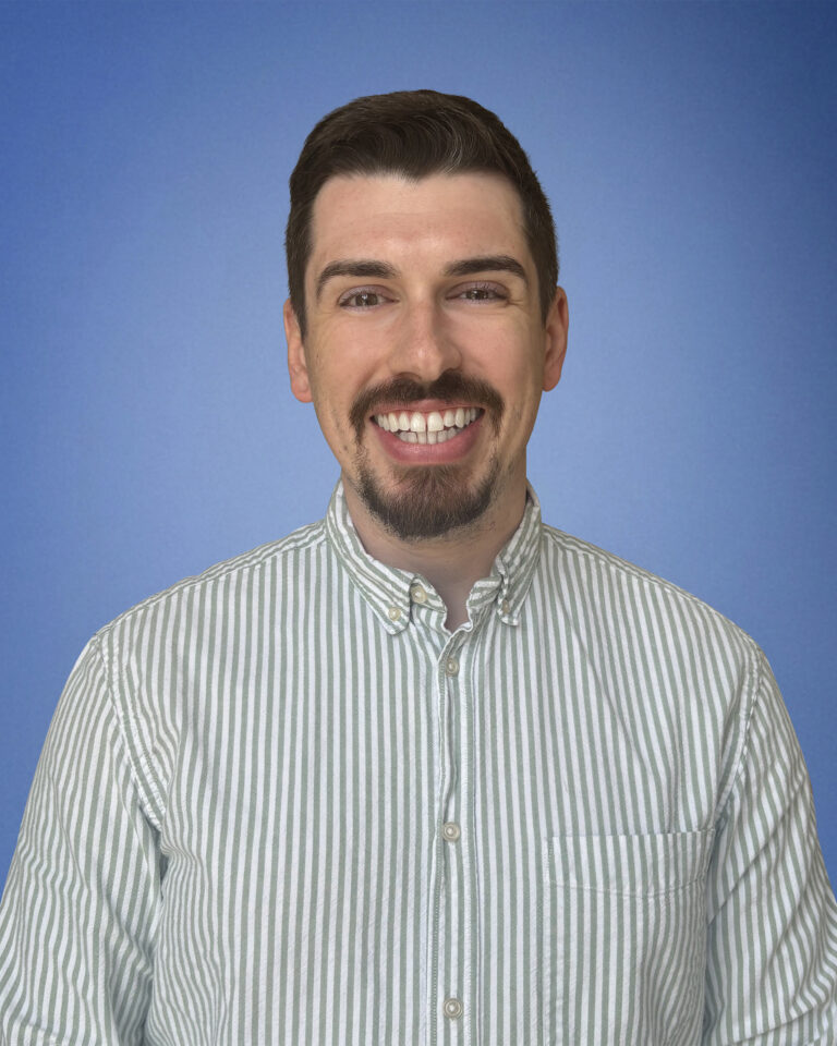 Smiling man with dark hair and a goatee wearing a striped button-up shirt against a blue background.