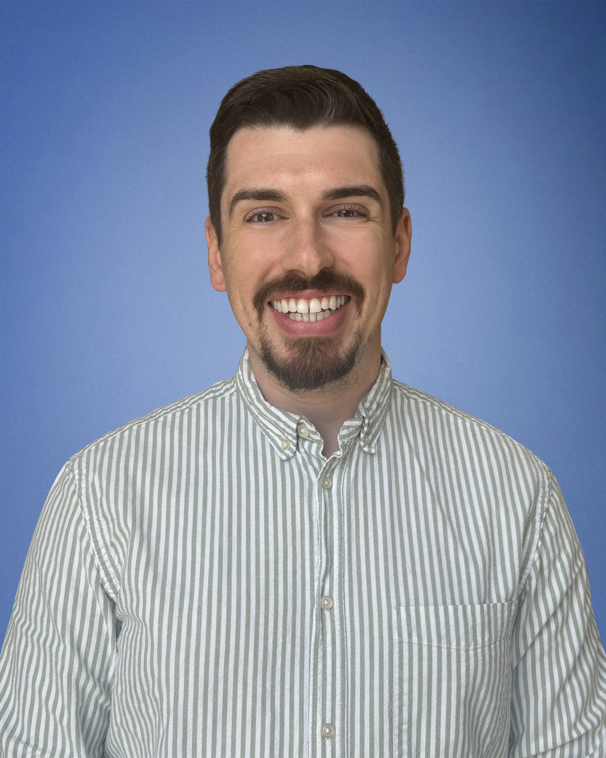 Smiling man with dark hair and a goatee wearing a striped button-up shirt against a blue background.