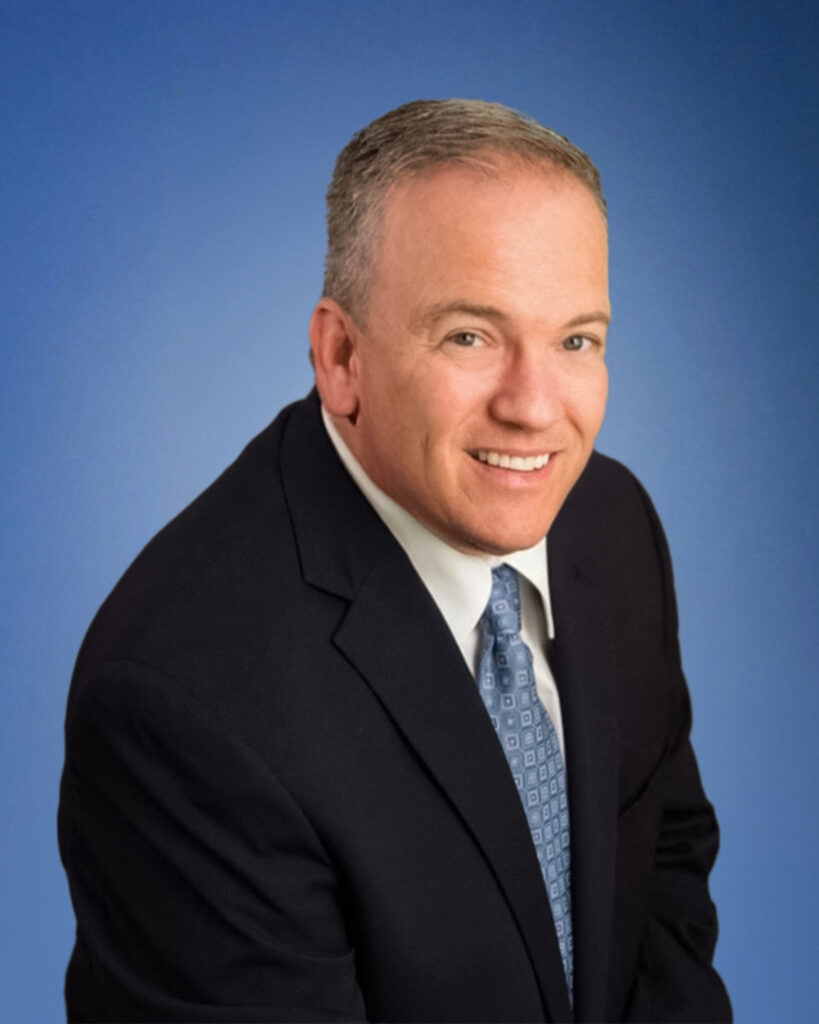 Smiling man in a dark suit and light blue tie against a blue backdrop, cropped head-and-shoulders portrait.