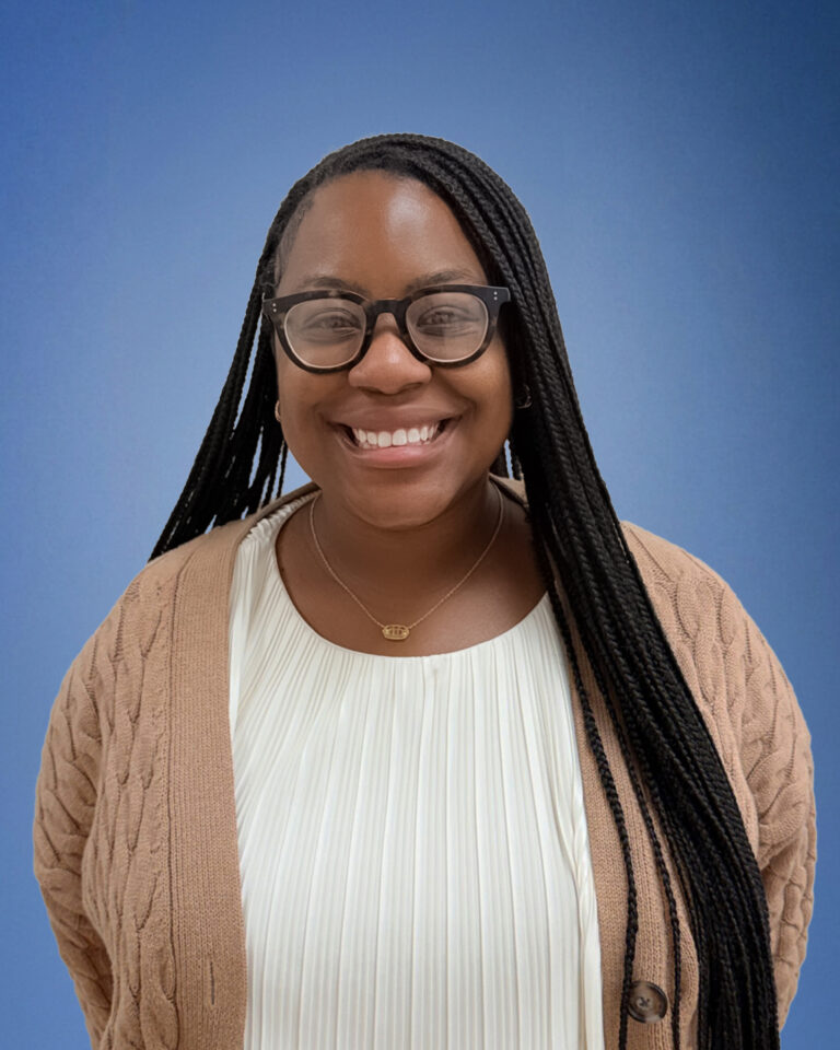 Smiling Black woman with long braided hair and black-framed glasses, wearing a beige cardigan over a white pleated top, against a blue background.