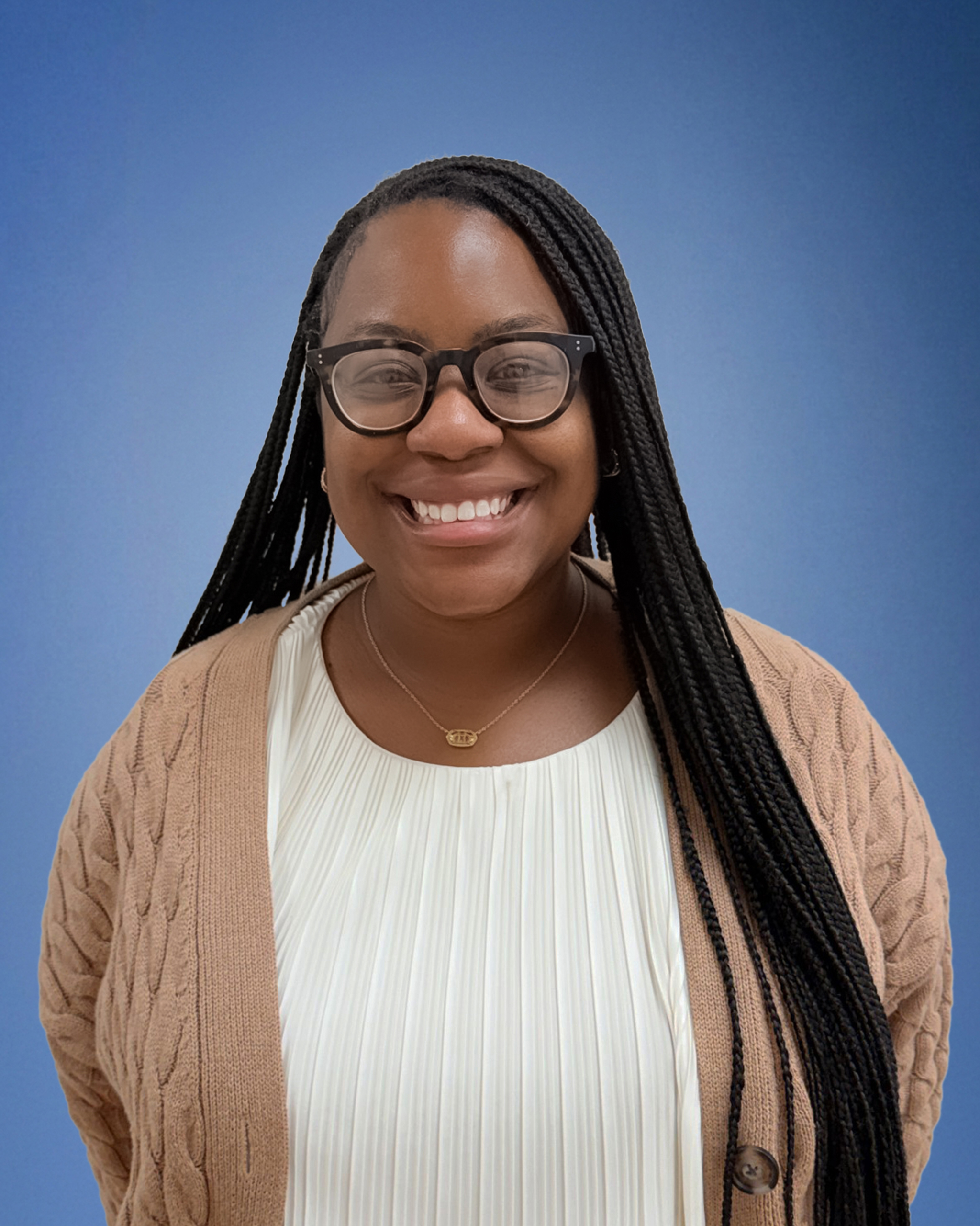 Smiling Black woman with long braided hair and black-framed glasses, wearing a beige cardigan over a white pleated top, against a blue background.
